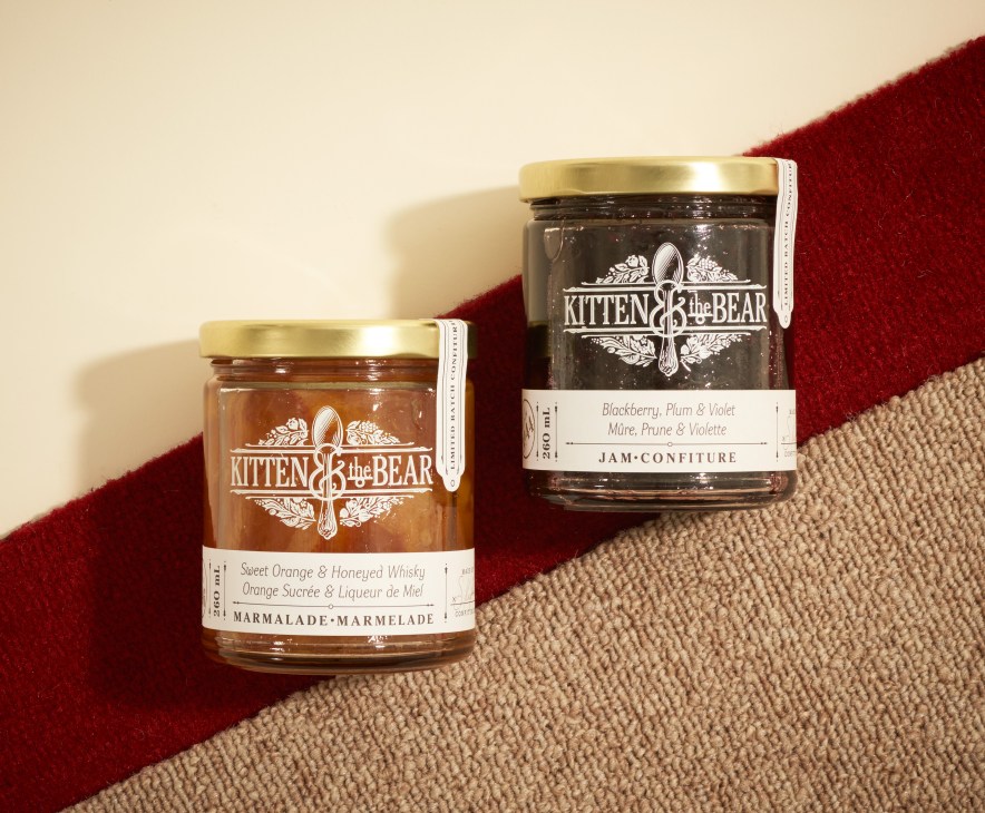 Two glass jars of Kitten & the Bear preserves with gold lids on a beige and burgundy striped surface