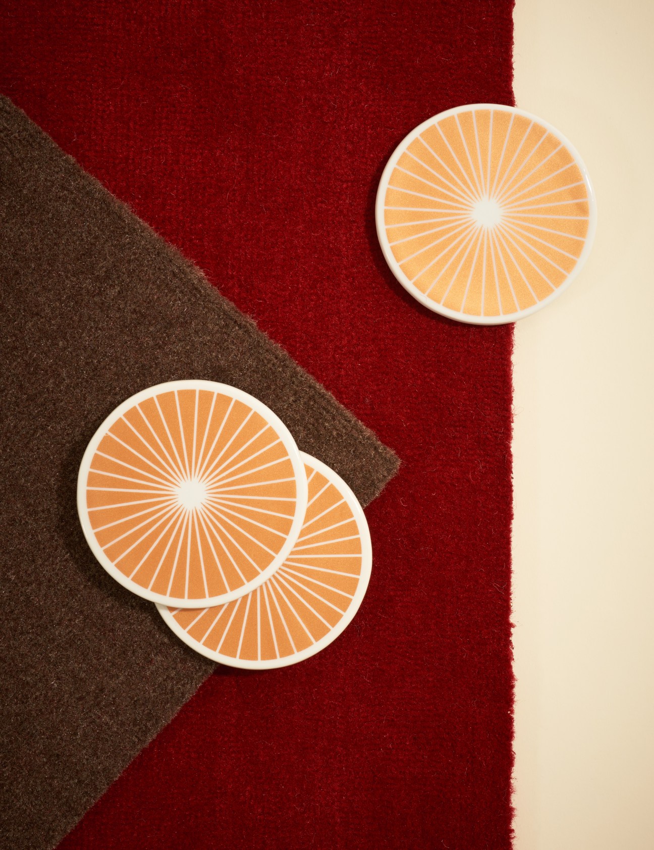Three orange ceramic coasters with radial spoke patterns displayed on colorful felt surfaces