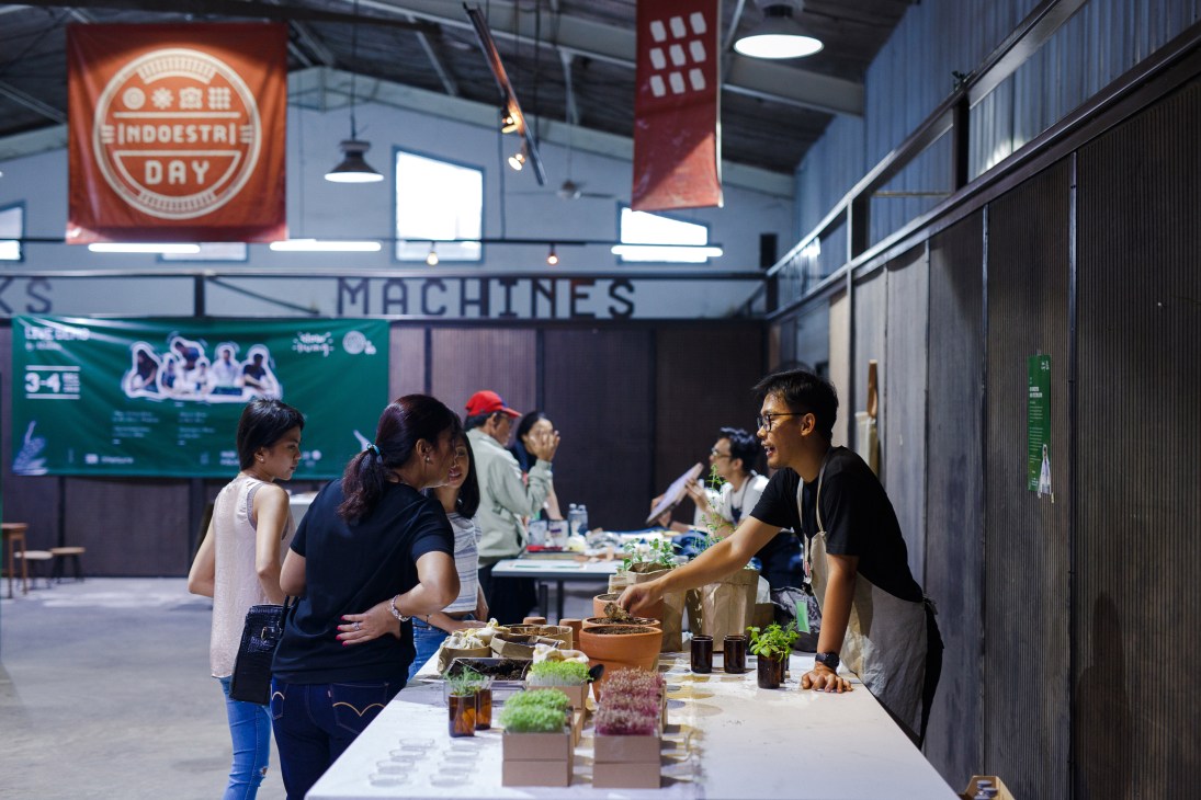 People browsing plants and products at Indoestri workshop with neon sign overhead.