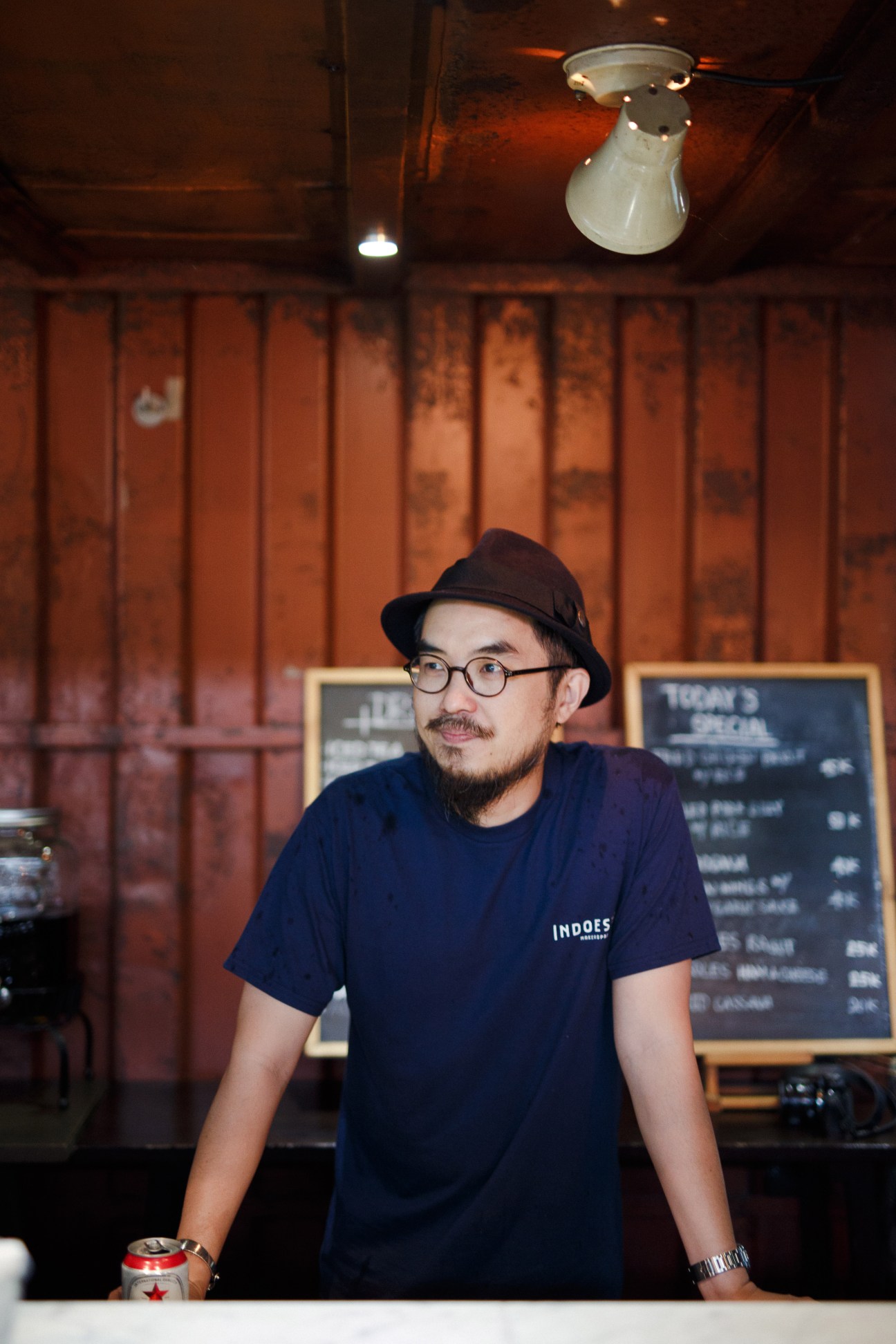 Man wearing glasses and black hat stands in front of rustic red-brown corrugated metal wall at Indoestri workspace.