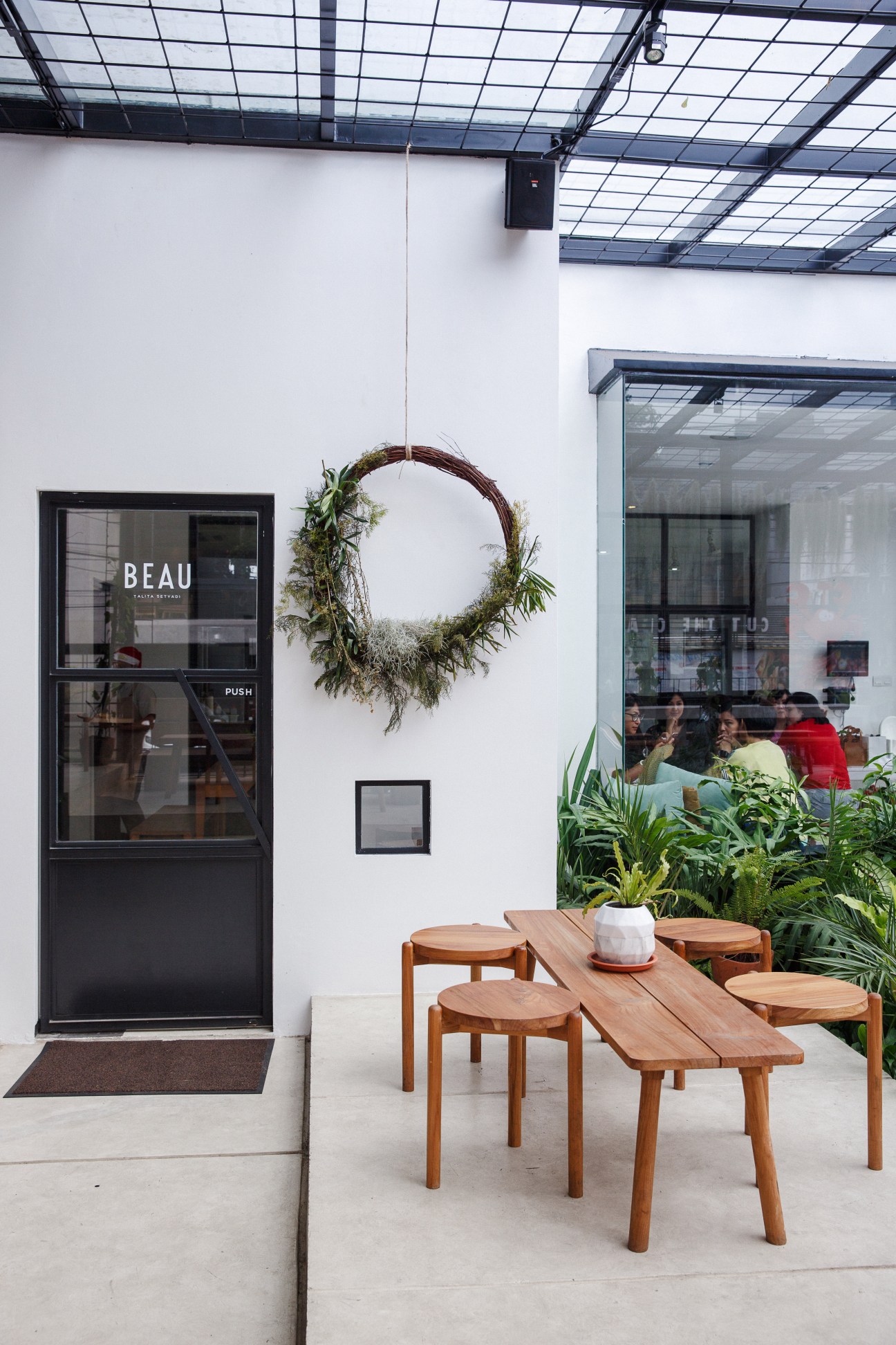 Beau café storefront with wooden furniture, potted plants, and a decorative wreath on white wall.