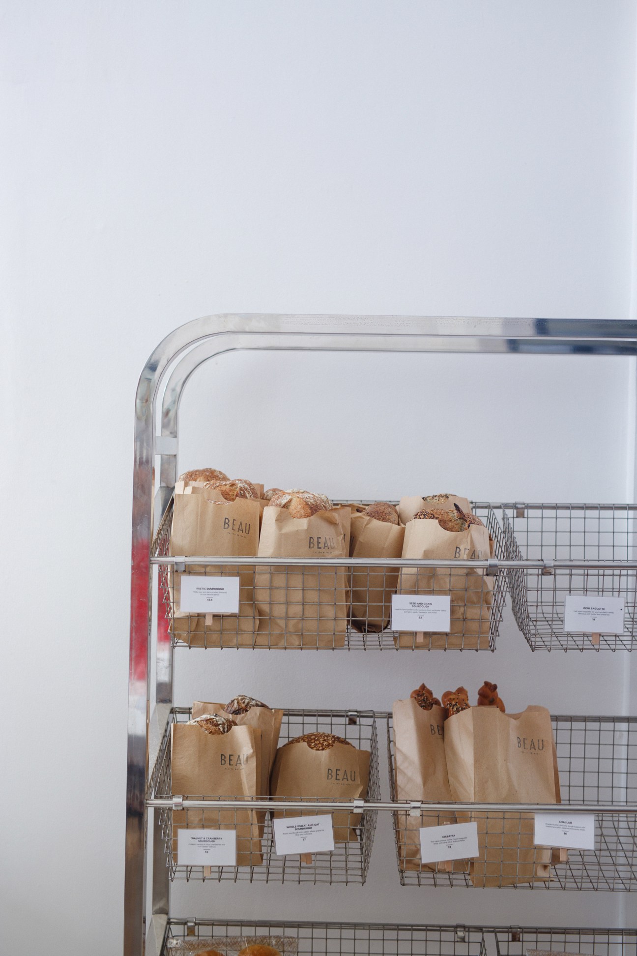 Beau bakery display shelf with paper bags of bread and pastries arranged on metal wire baskets.