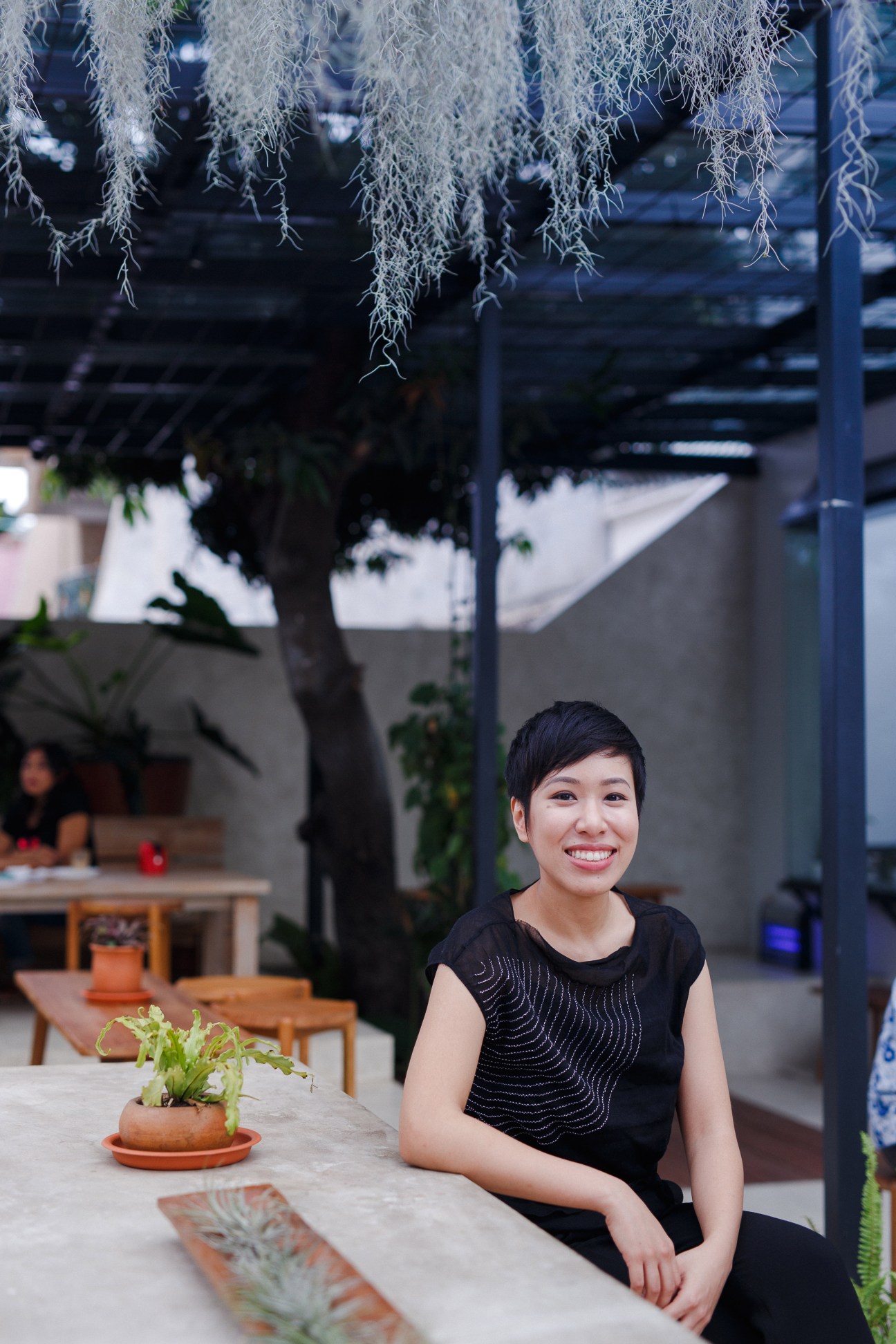 Talita Setyadi, founder of Beau bakery, smiles while sitting at a table in an outdoor courtyard with hanging plants overhead.