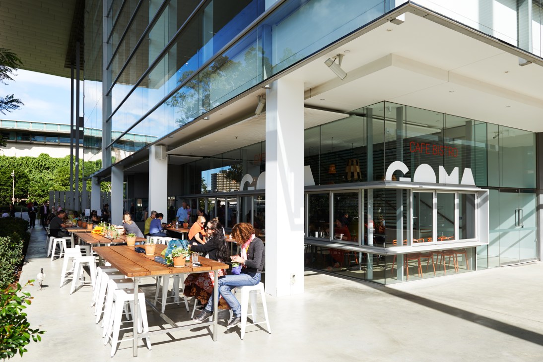 Visitors seated at wooden tables under a large cantilevered roof at Goma's outdoor café overlooking gardens.