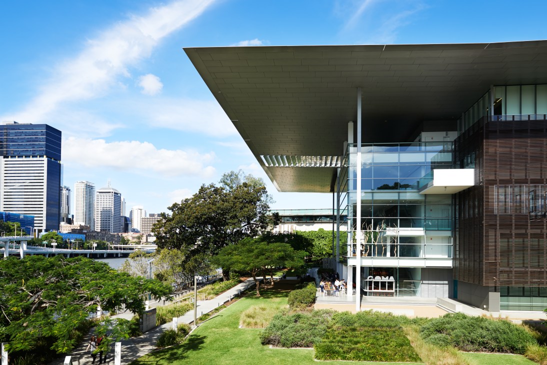 Gallery of Modern Art building with cantilevered roof overlooking gardens and Brisbane River with city skyline.