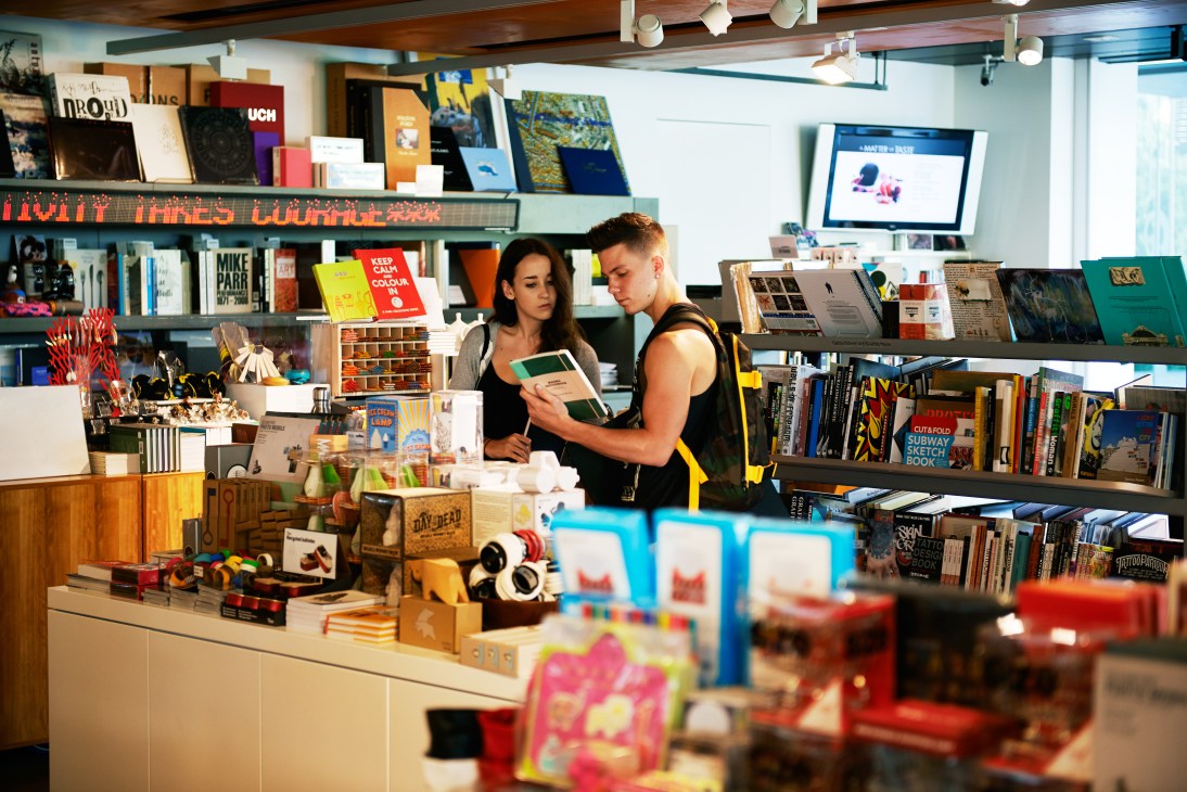 Two customers browse books and merchandise at Goma's gallery shop with shelves of art books and products displayed behind them.