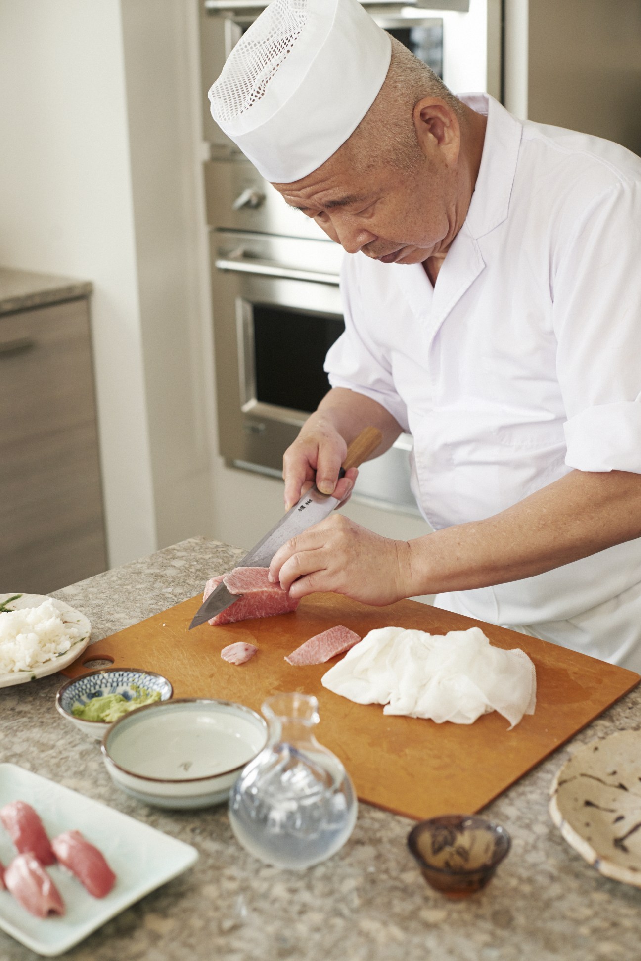 Toshihiro Uezu in white chef's attire and hat carefully slicing raw fish on a wooden cutting board.