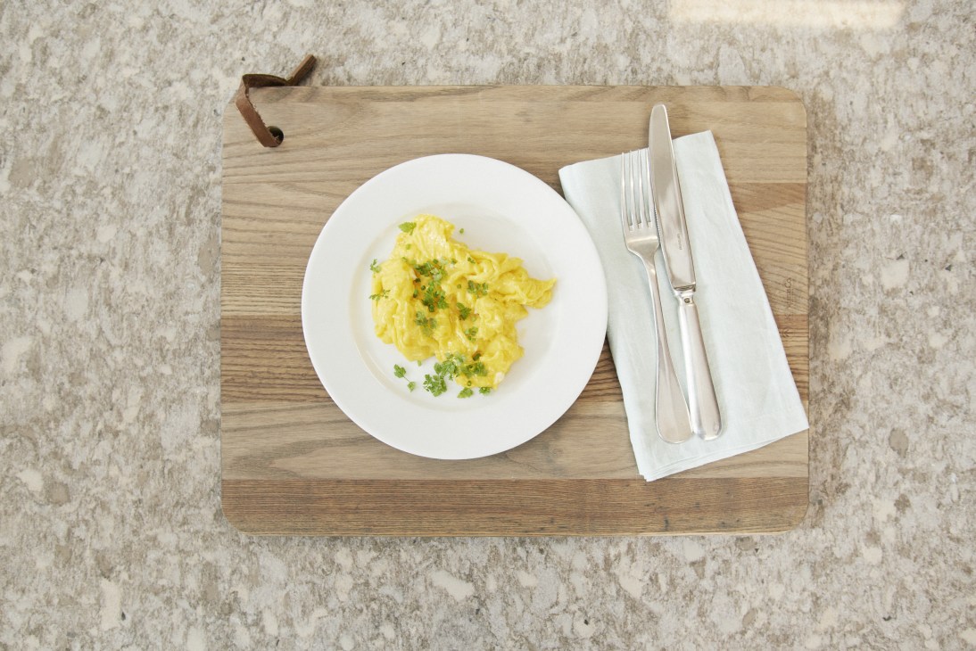 Fluffy scrambled eggs garnished with chives on a white plate with cutlery on a wooden board.