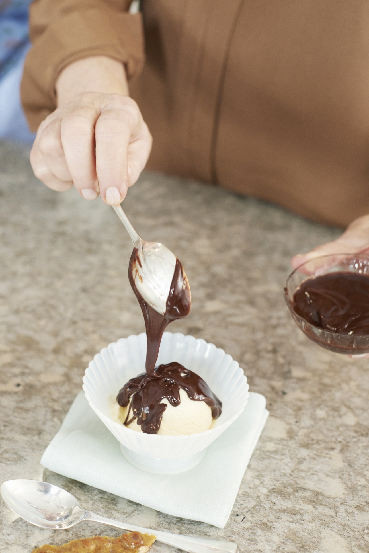 Hand drizzling hot fudge over vanilla ice cream in a white bowl.