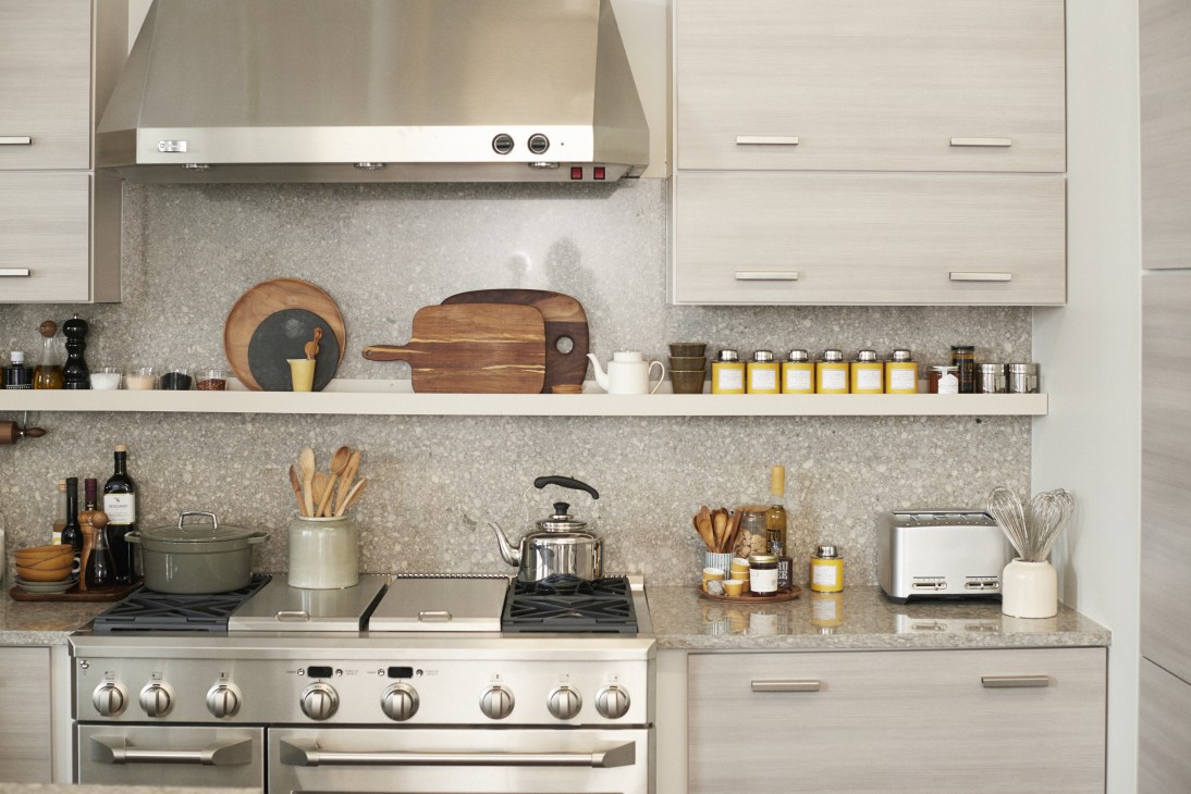 Modern test kitchen with cream cabinets, stainless steel range, open shelving displaying wooden cutting boards and yellow glass jars.