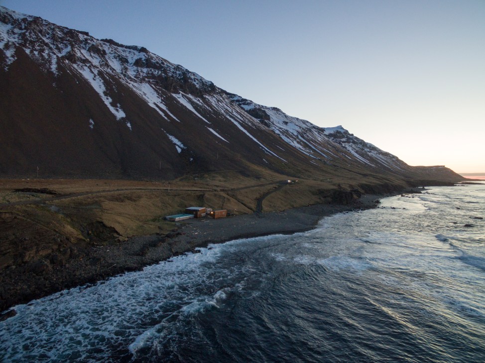 Krossneslaug geothermal pool nestled on black volcanic pebbles below a snow-capped mountain in Arctic Iceland.