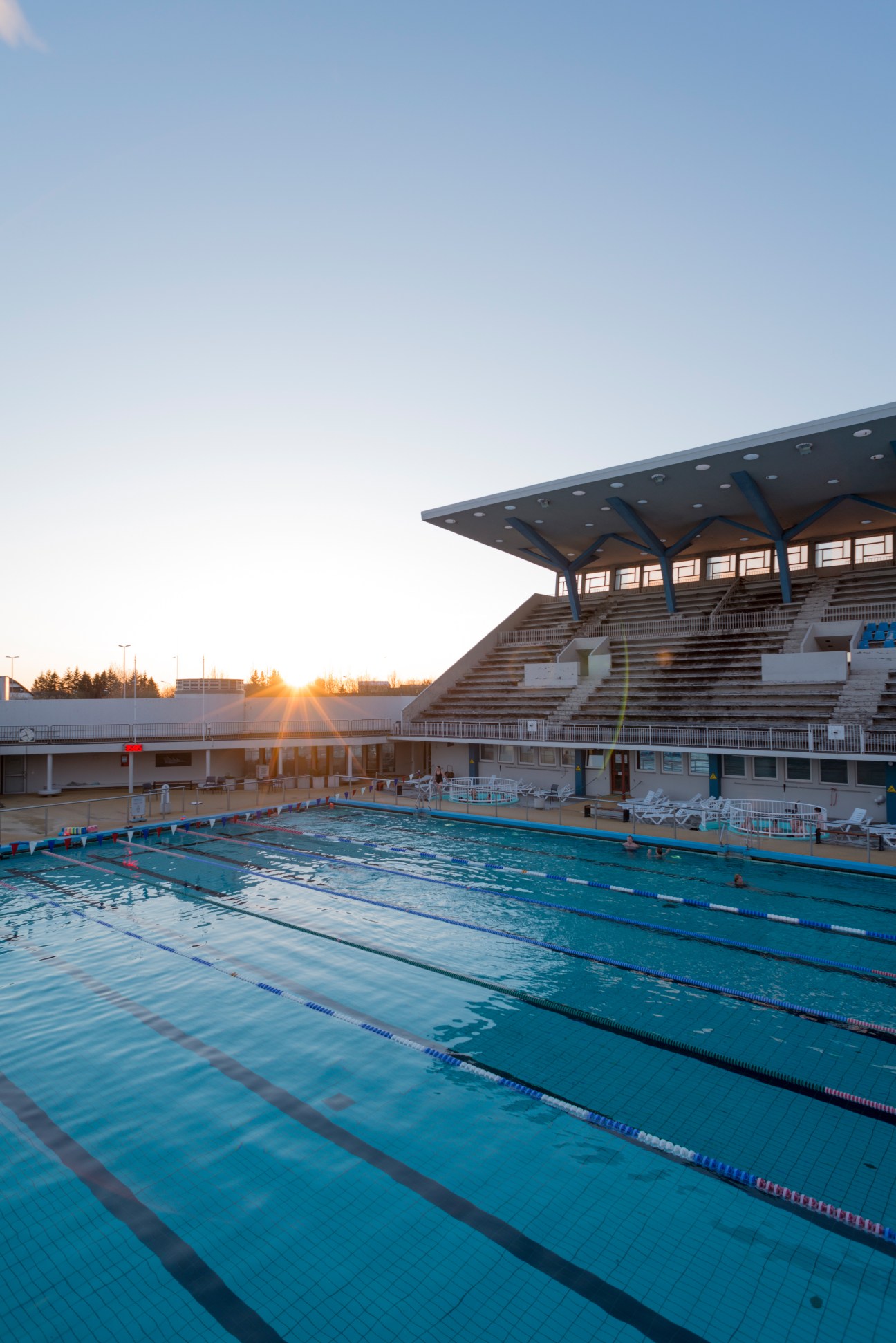 Empty outdoor Olympic swimming pool with lane dividers at sunrise or sunset, with modern stadium seating and facility building.
