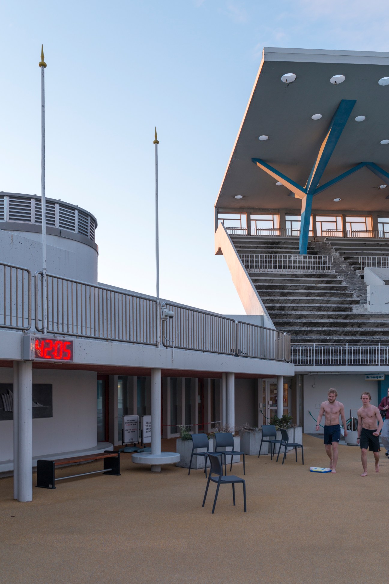 Modern poolside architecture with blue structural elements and two shirtless swimmers leaving at closing time