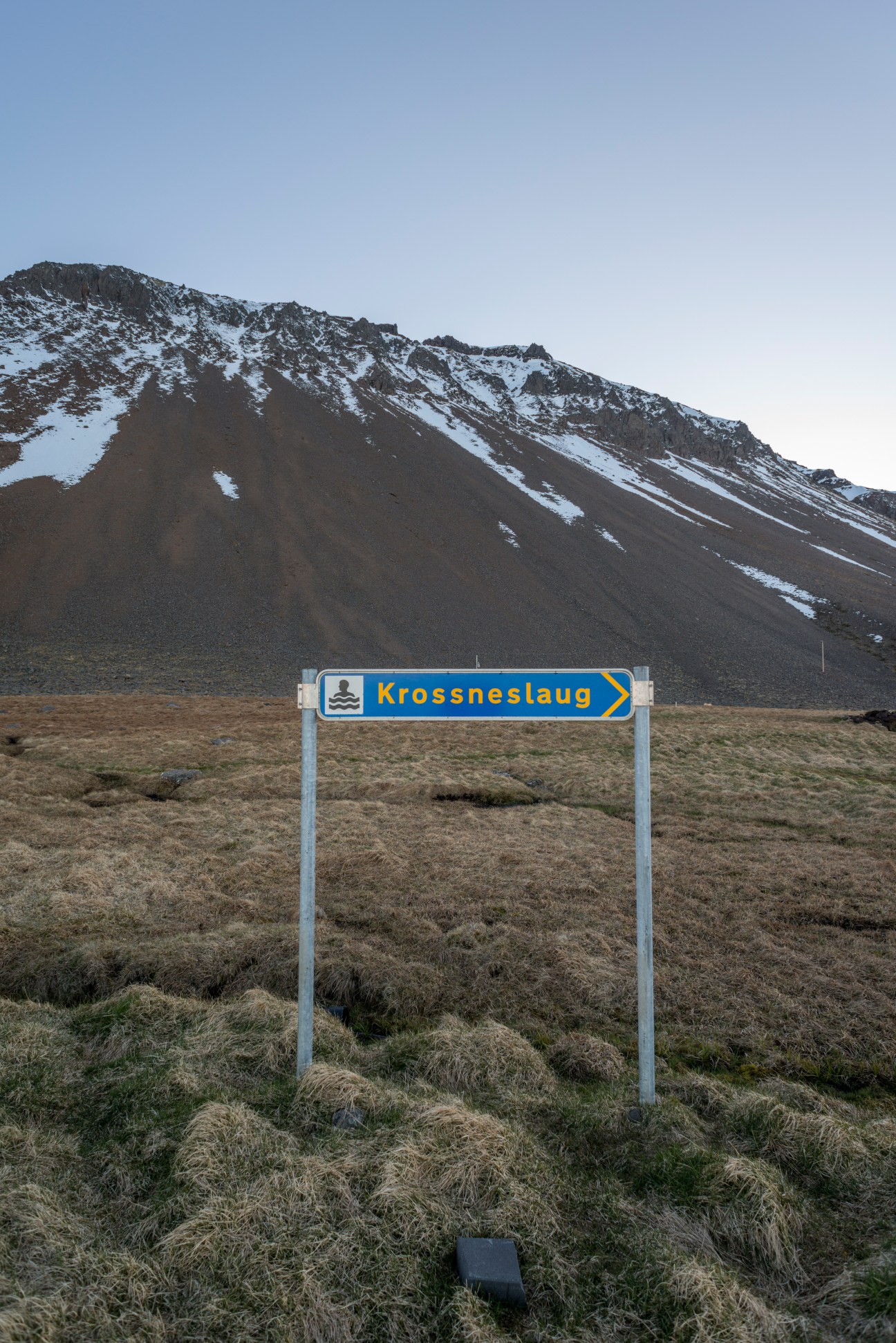 A blue and yellow directional sign for Krossneslaug pointing toward a snow-capped volcanic mountain in Iceland.