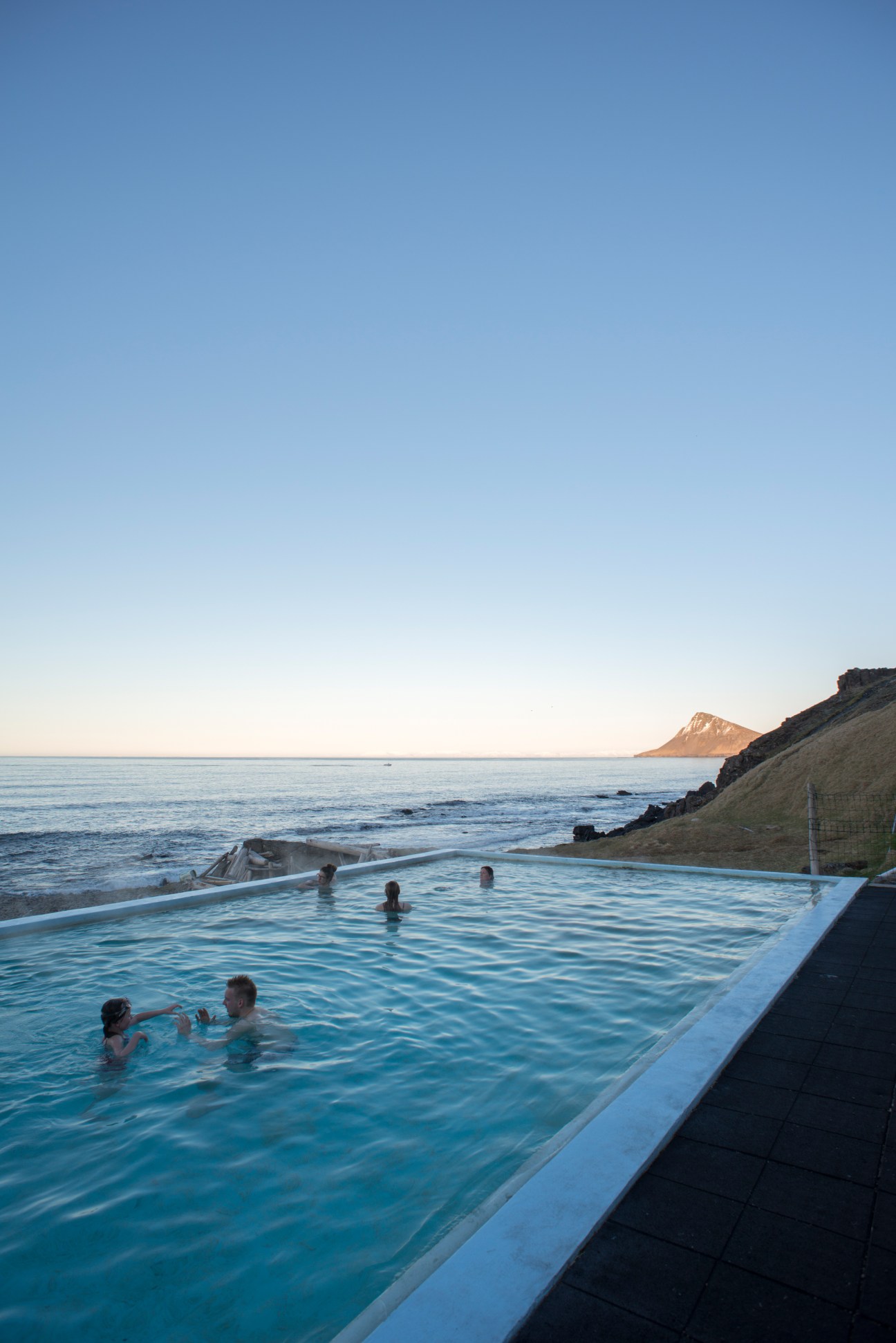 Bathers enjoying the warm geothermal pool at Krossneslaug in Iceland with sea and snow-capped mountains in the background at dusk.