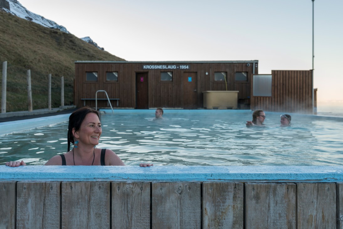 Woman relaxing in the warm waters of Krossneslaug pool in Iceland with other bathers visible in the background.