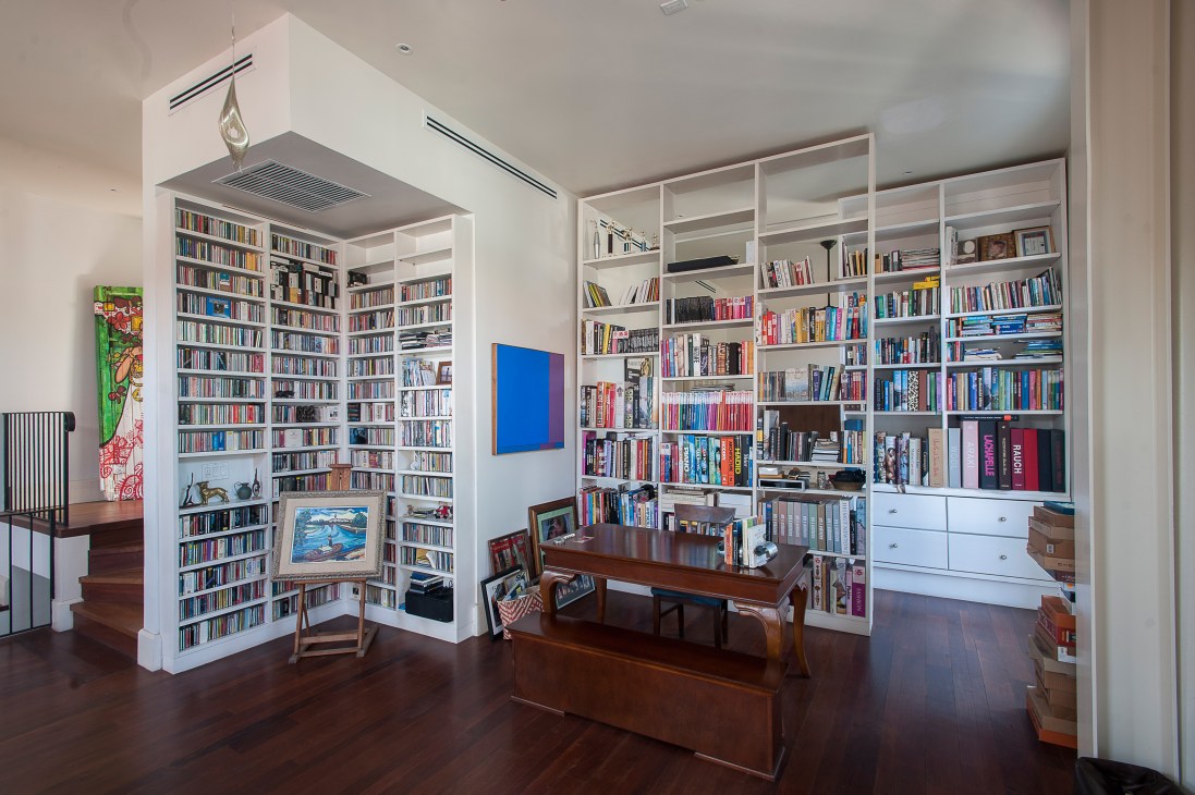 A modern library room with floor-to-ceiling white shelving filled with books, a wooden desk, and colorful artwork on white walls.