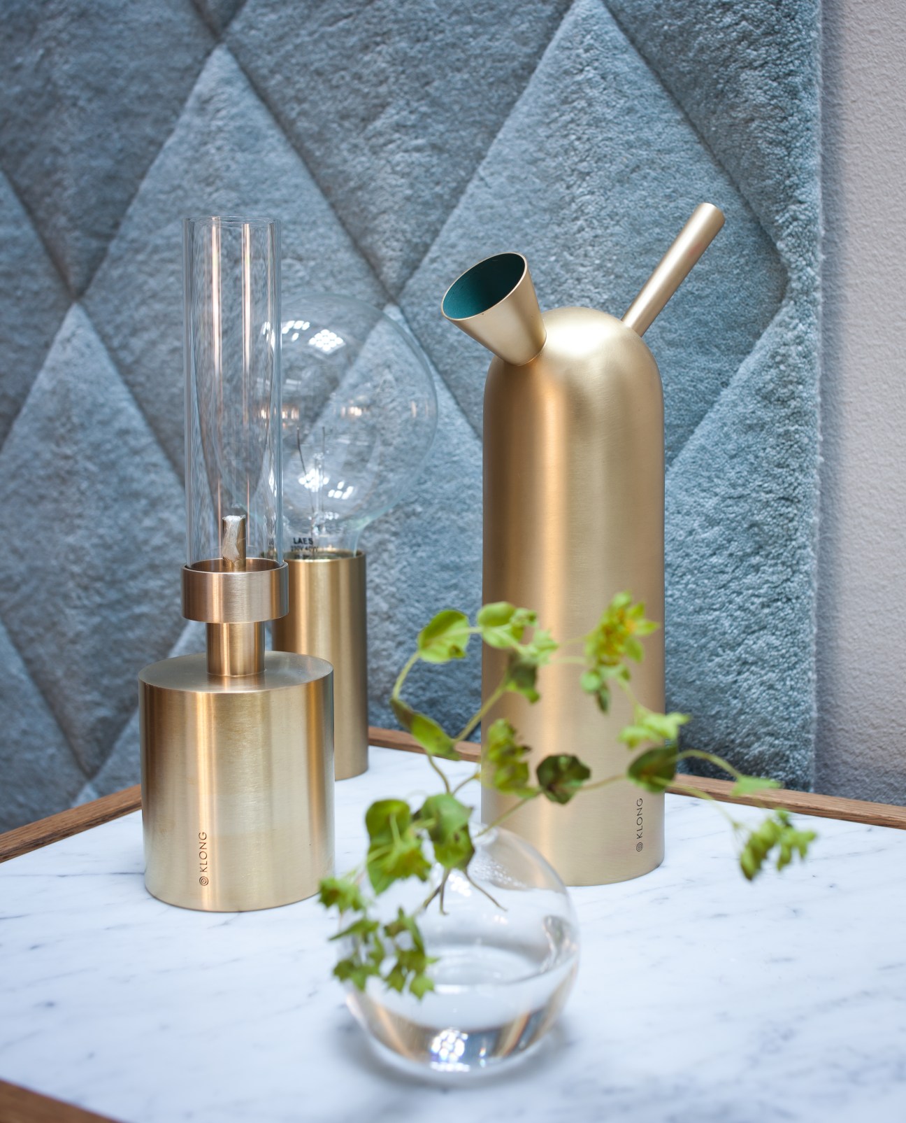 Broberg & Ridderstråle's brass oil lamp and water bottle displayed on marble with potted greenery against textured blue wall.