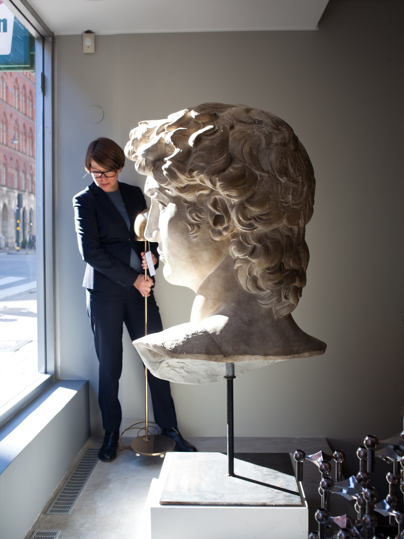 Woman in dark suit standing beside a large classical stone bust displayed in a bright window.