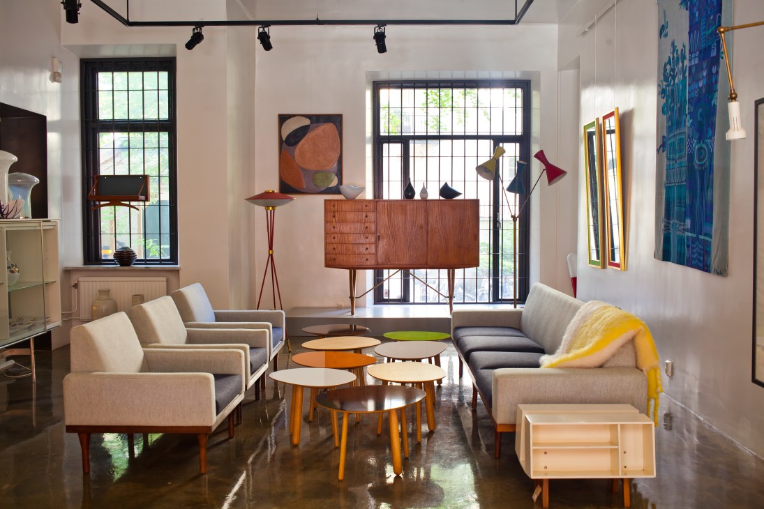 Mid-century modern furniture showroom with beige armchairs, wooden credenza, colorful nesting tables, and pendant lamps.