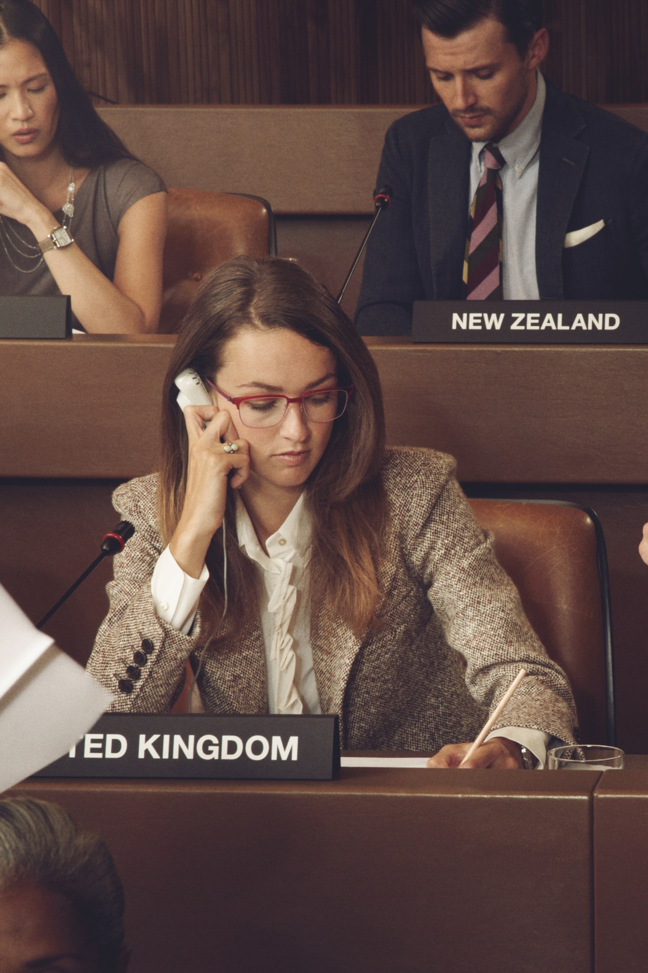 Woman diplomat in tweed blazer and glasses at UN assembly desk with microphone.