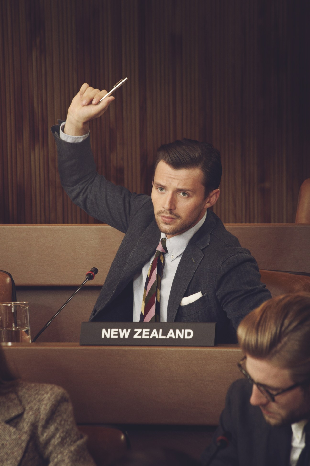 Model representing New Zealand at UN General Assembly wearing dark suit and striped tie, raising pen at conference desk.