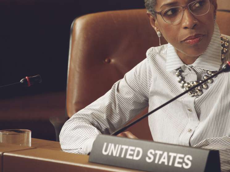 Model seated at diplomatic table wearing striped shirt and statement necklace with United States nameplate.
