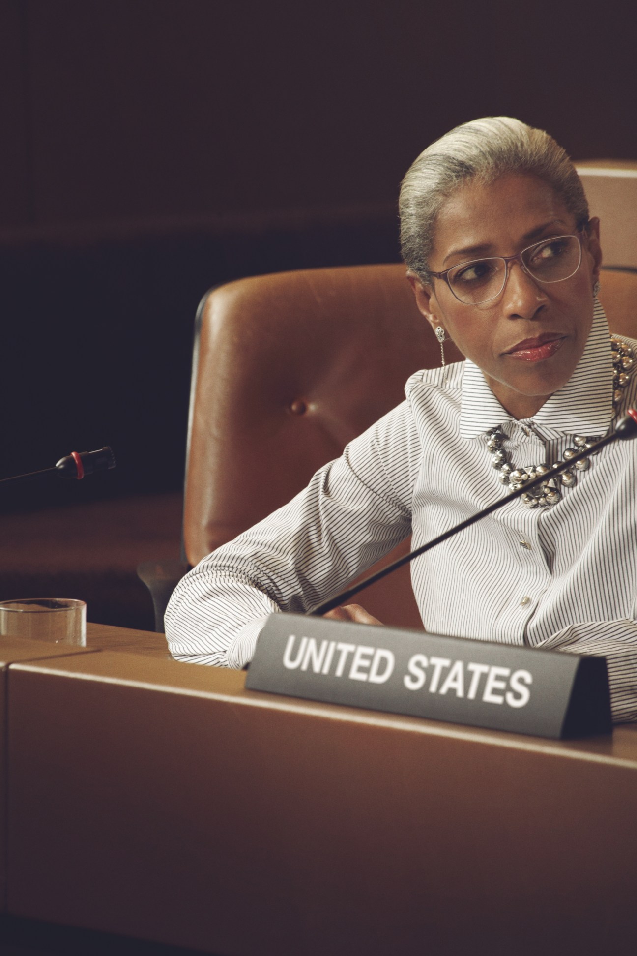 Model seated at diplomatic table wearing striped shirt and statement necklace with United States nameplate.