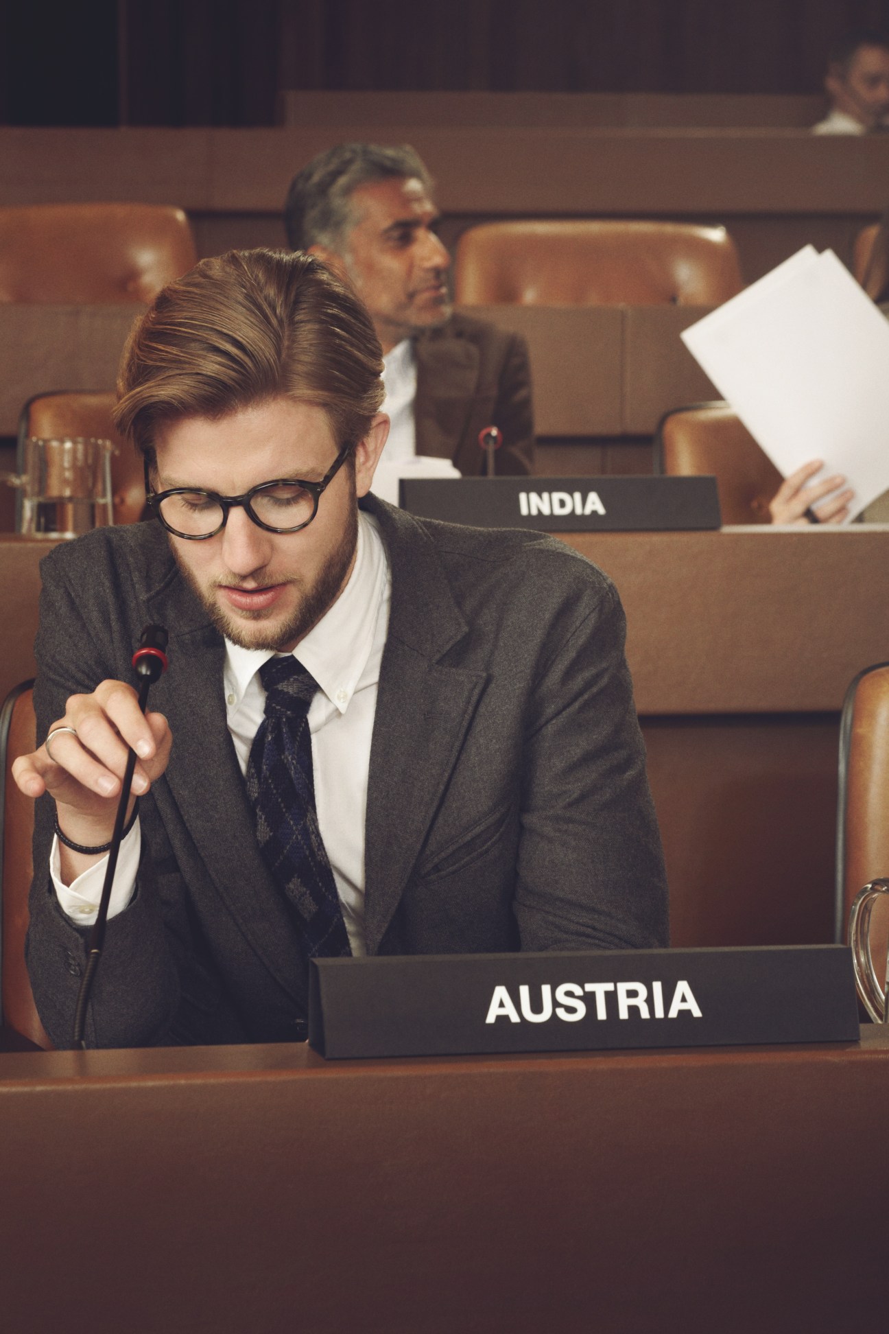 Male model in grey suit and glasses sitting at UN General Assembly desk labeled Austria, holding microphone.