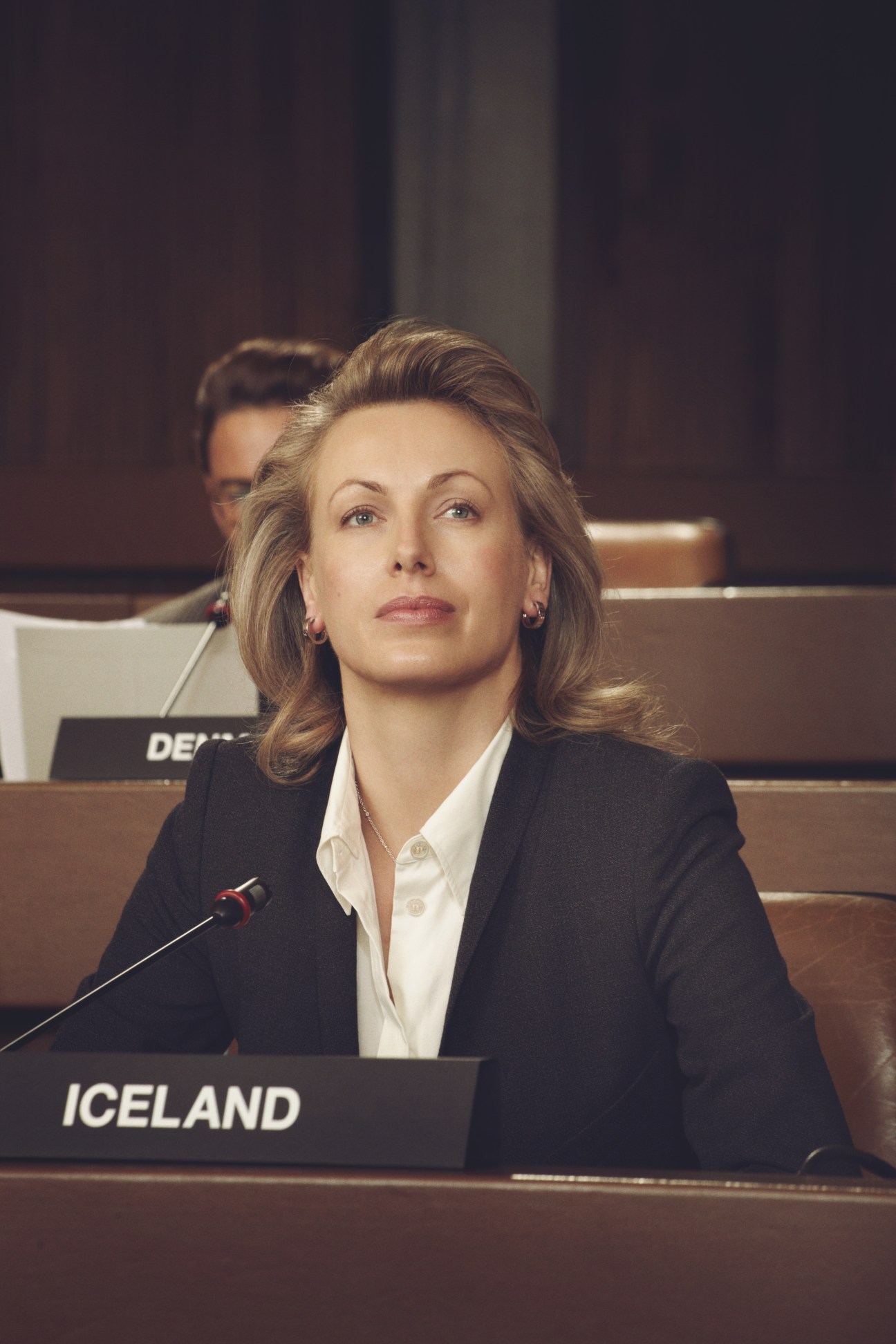 Female model dressed as Iceland delegate seated at UN General Assembly with nameplate and microphone.