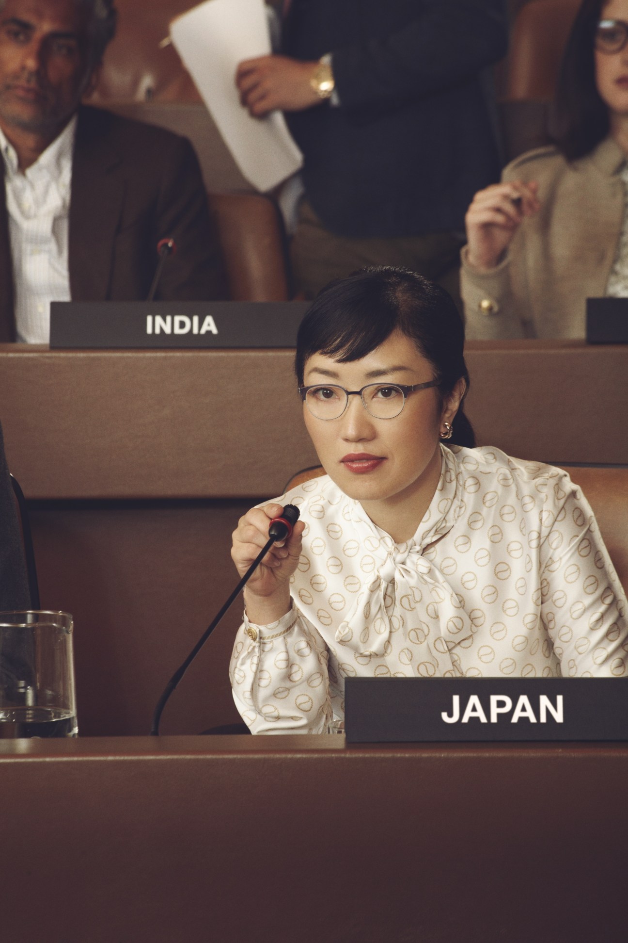 Model in patterned cream blouse seated at UN assembly desk labeled Japan, holding microphone.