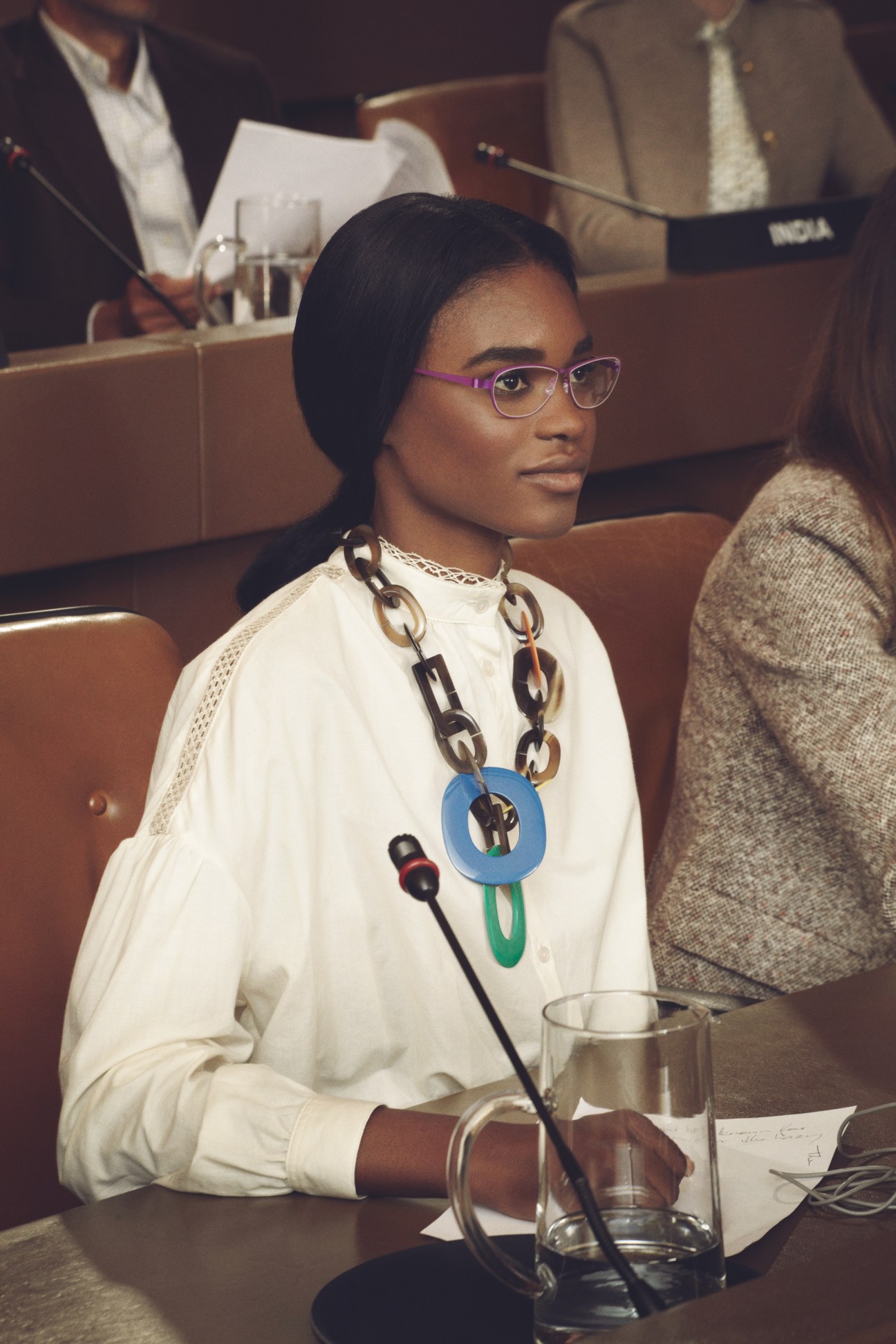 Model wearing white shirt and chain necklace seated at UN General Assembly desk labeled India.
