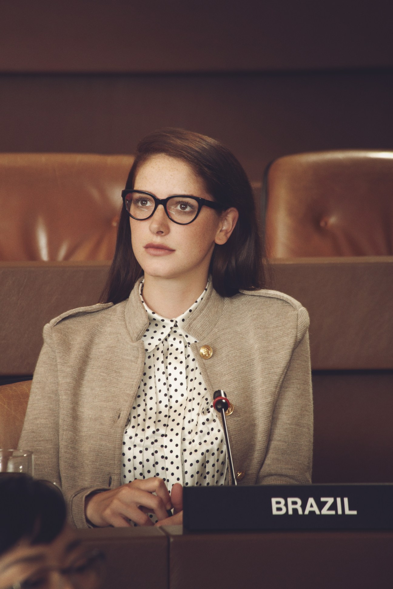 Model wearing beige blazer and polka-dot blouse with glasses, seated at UN General Assembly desk labeled Brazil.