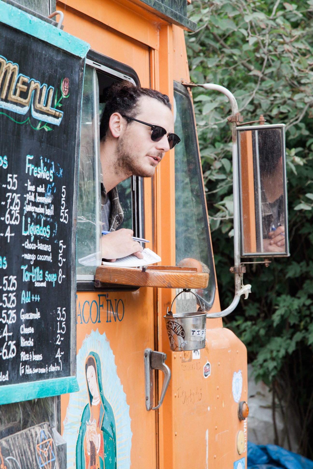 Man in sunglasses serving from Tacofino's bright orange food truck window with menu board visible