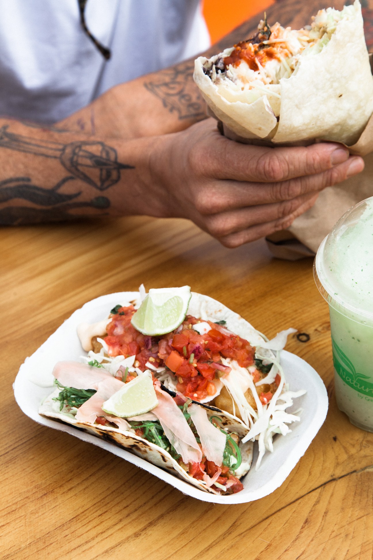 Tattooed hands holding a burrito with plate of tacos and green drink on wooden table at Tacofino