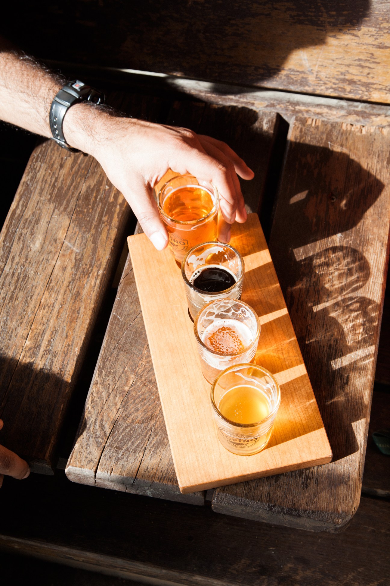 Hand reaching for beer flight of four different colored brews on wooden tasting board