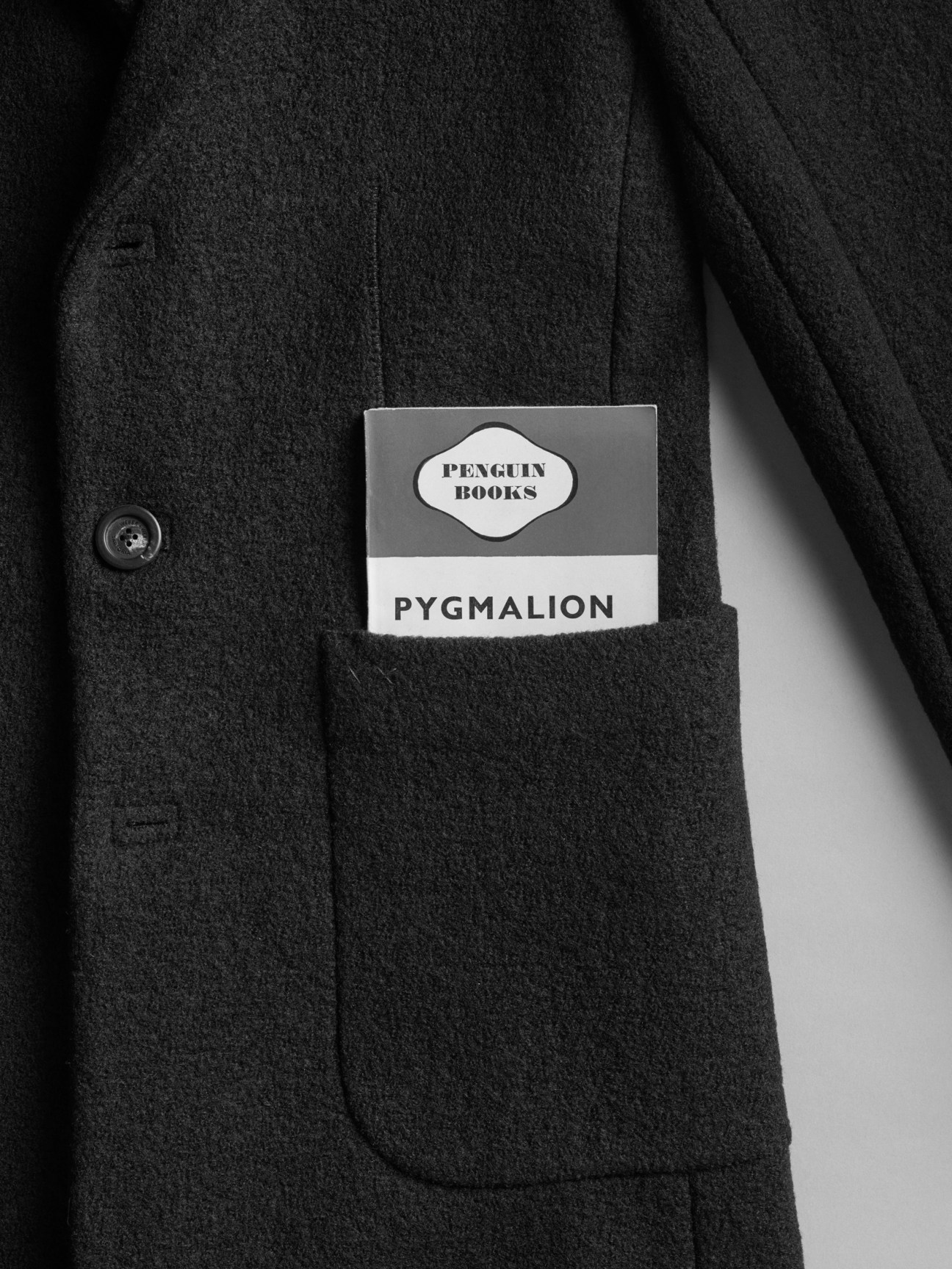 Close-up of a dark wool coat pocket with a white Penguin Books Pygmalion bookmark or label inserted