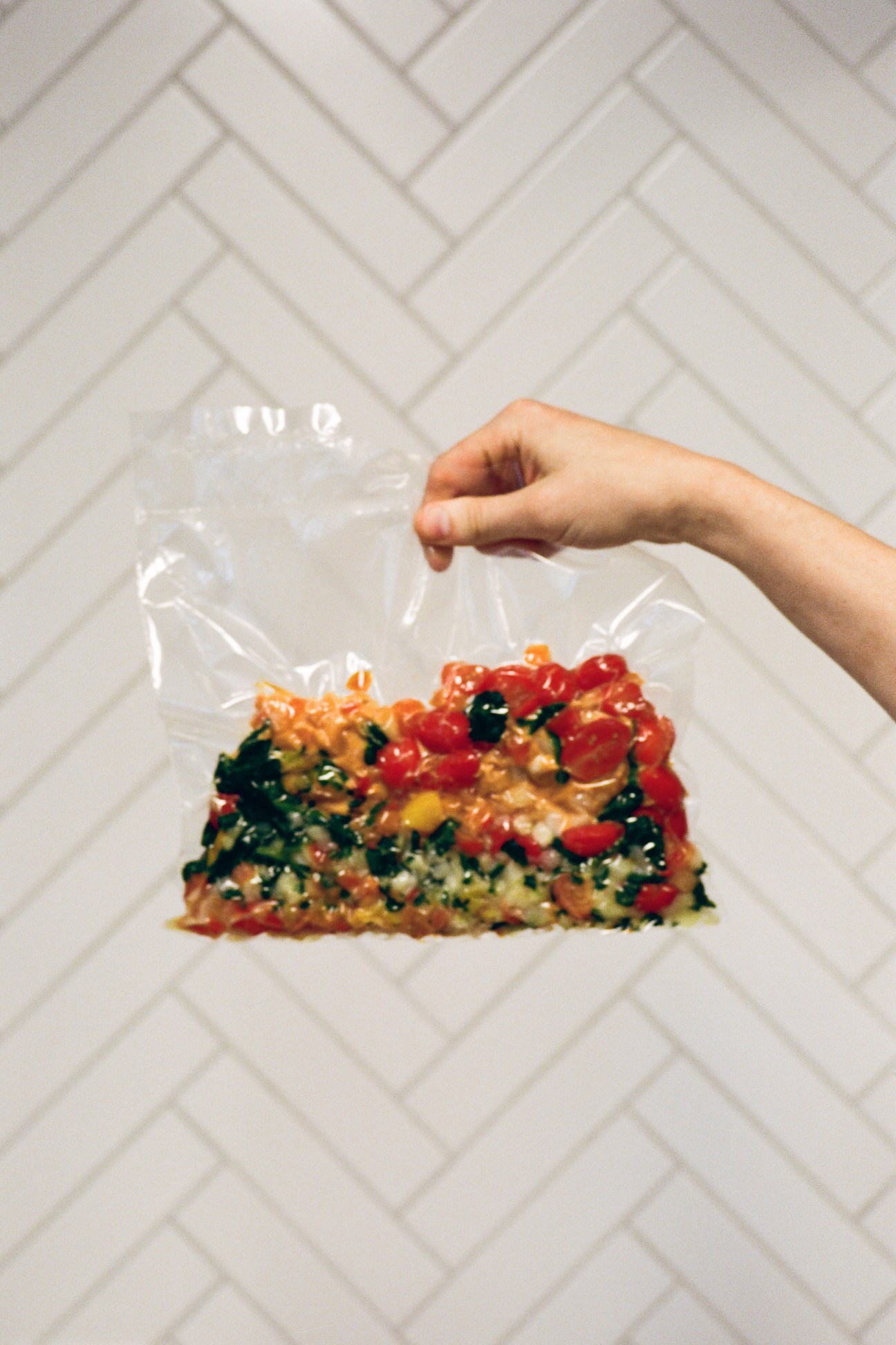 Hand holding plastic bag containing vacuum-packed food sample with vegetables against white herringbone tile background.