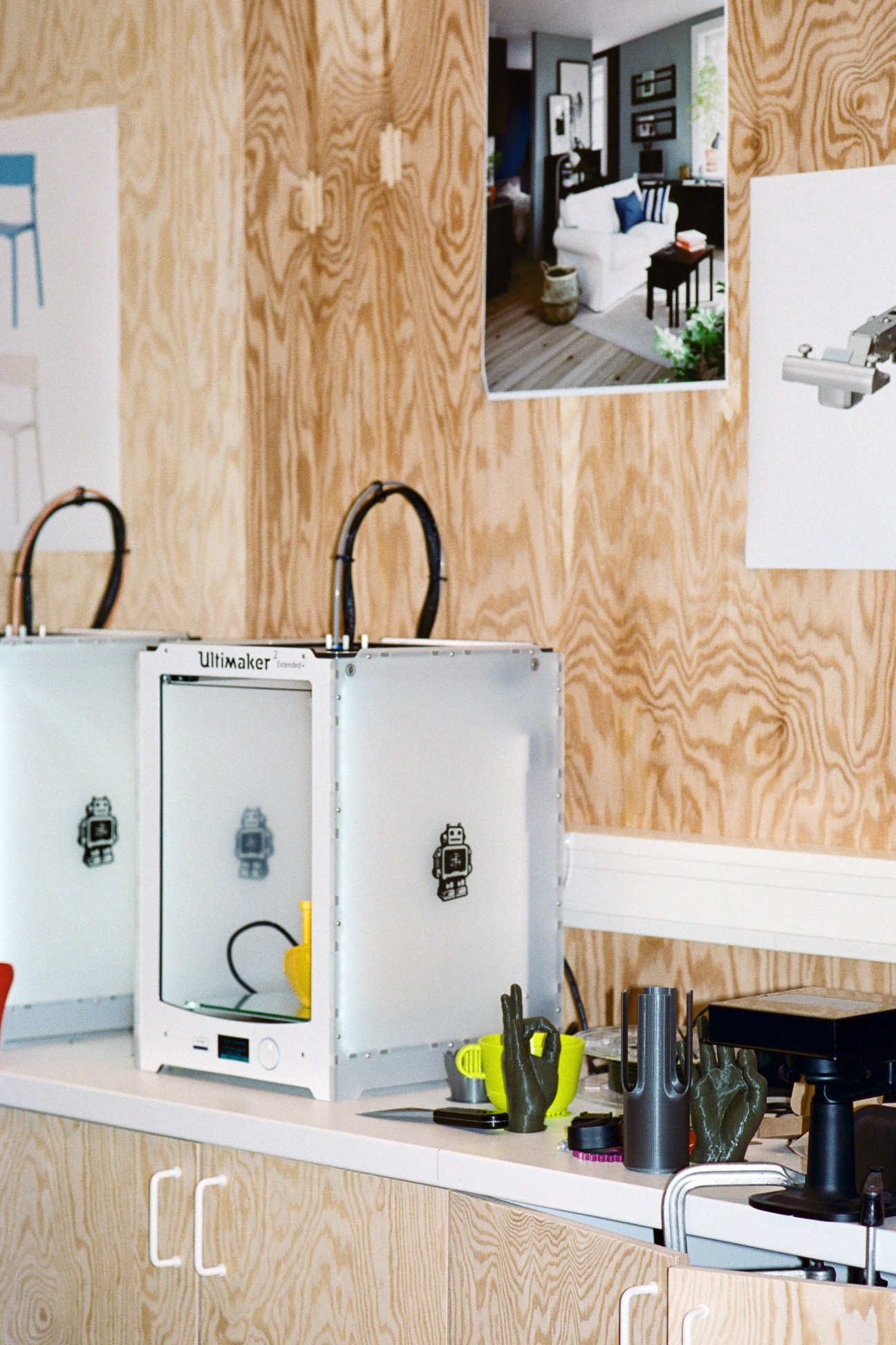 An Ultimaker 3D printer on a white desk against a plywood wall with interior design photos pinned above it.