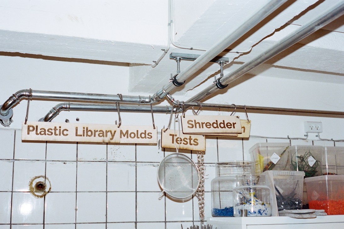 Laboratory workspace with labeled wooden signs for plastic library molds and shredder tests hanging from metal pipes above white tiled wall and containers of materials.