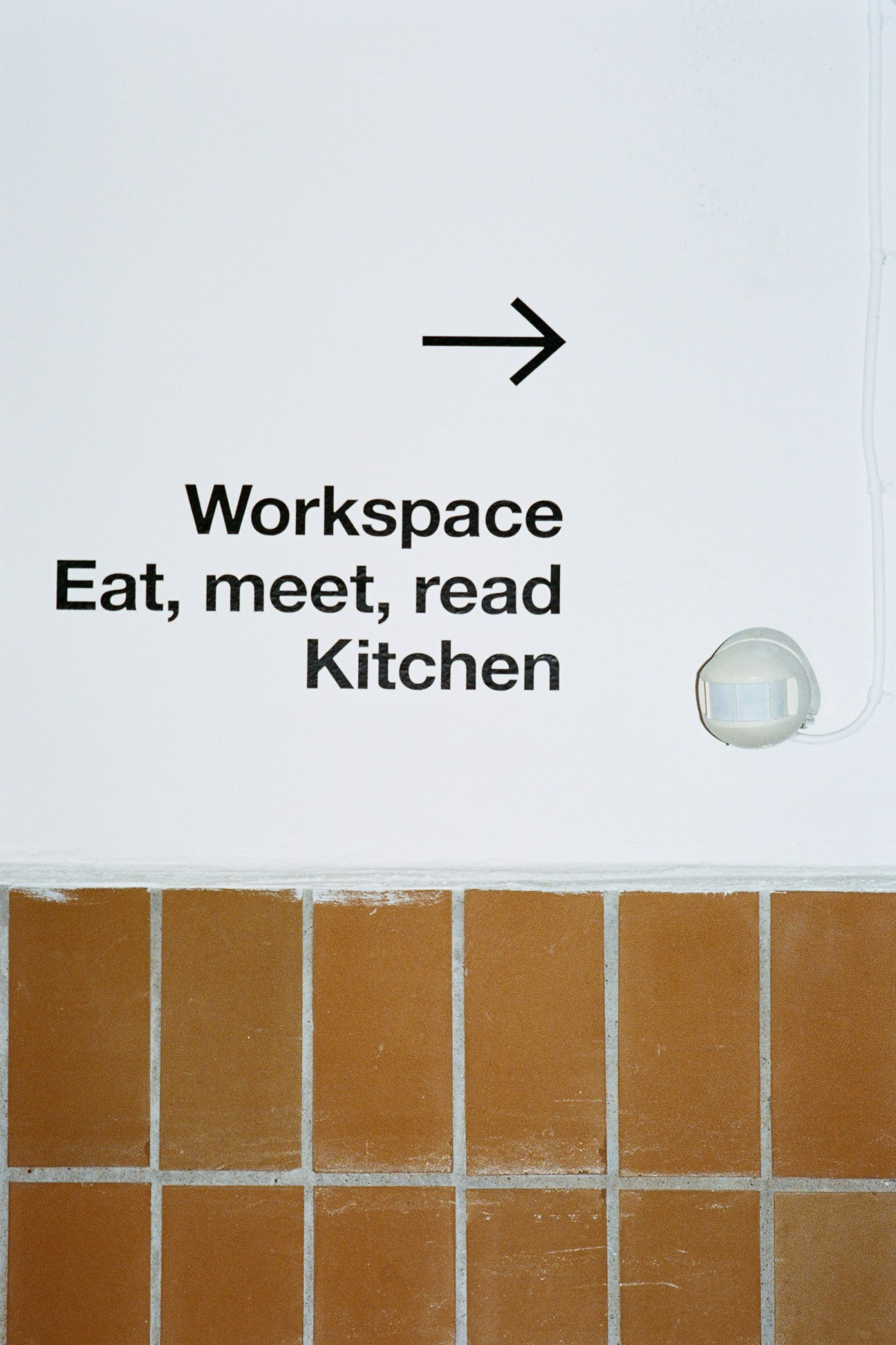 Interior wall signage reading 'Workspace Eat, meet, read Kitchen' with arrow and motion sensor on brick wall.