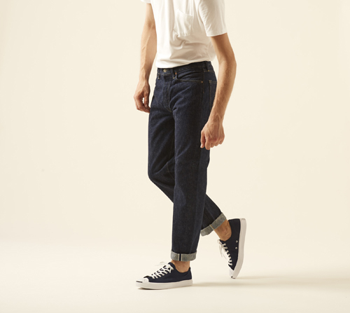 Model wearing Slim fit 107 jeans by OrSlow in dark indigo with cuffed hem and black sneakers.