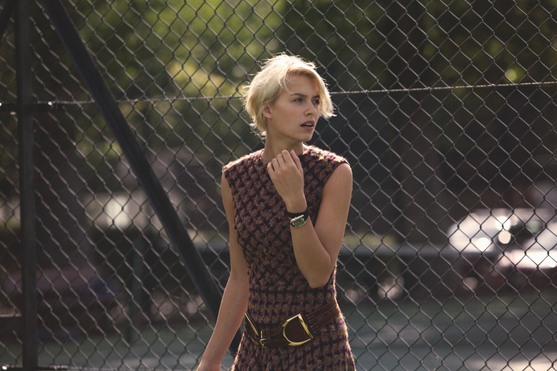 Woman in Oscar de la Renta dress and Ralph Lauren belt stands by tennis court fence.