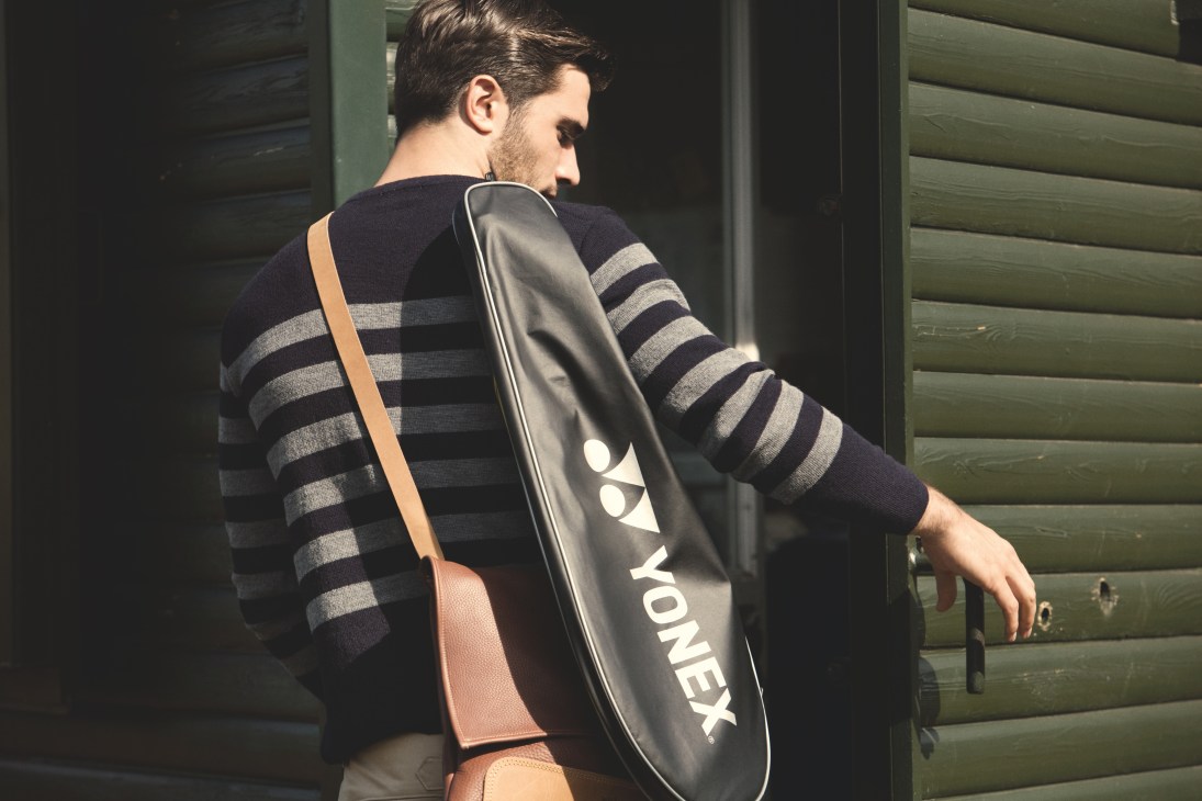 Man wearing striped jumper carrying Yonex racquet bag and shoulder bag against green shutters.
