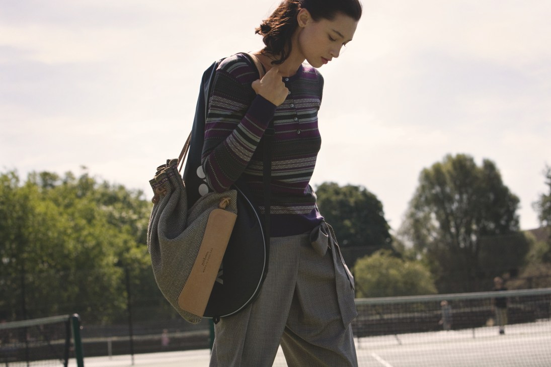Woman in striped jumper holding tennis racquet bag at Parliament Hill Fields courts.