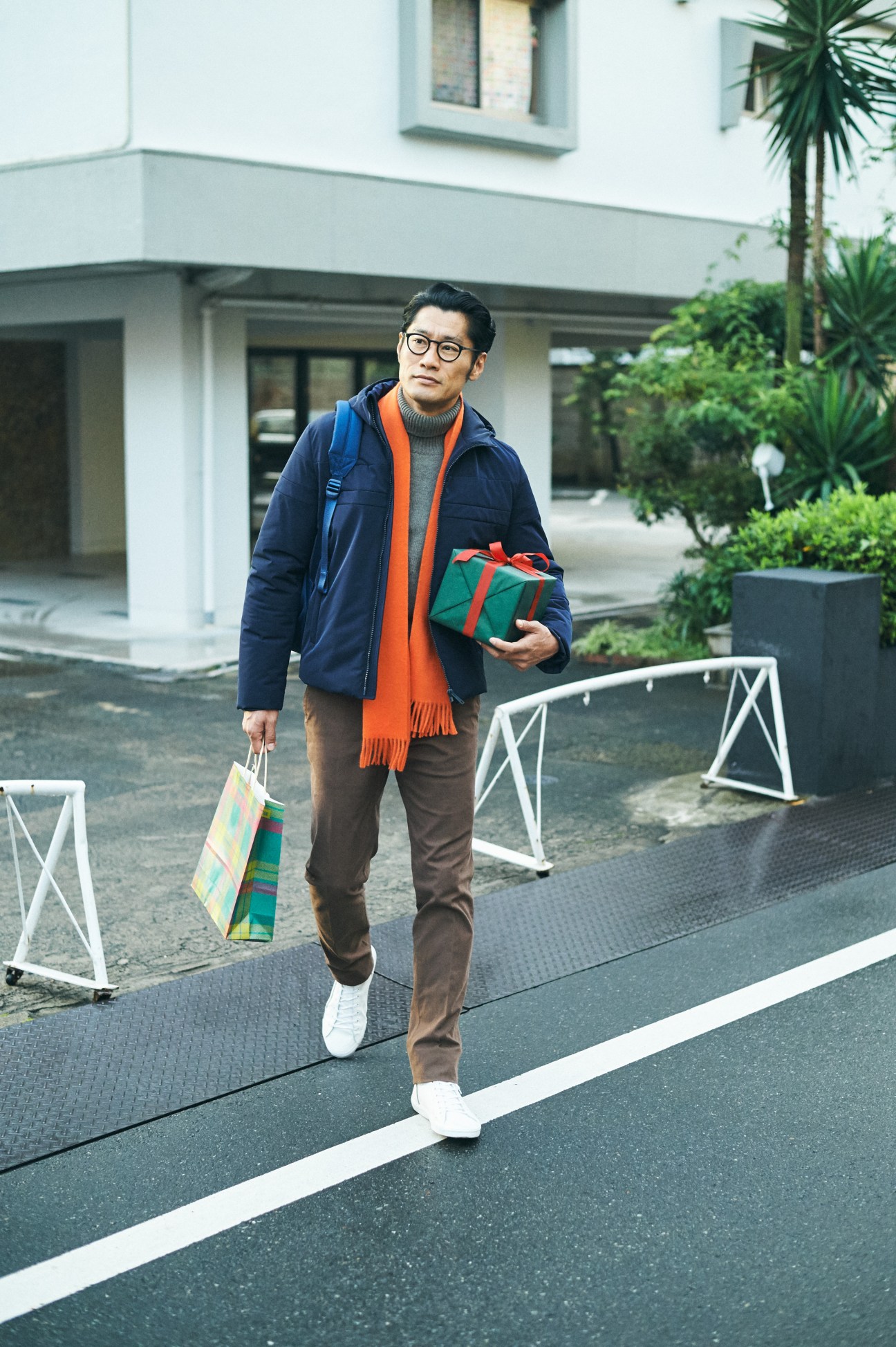 Man in navy Prada down jacket and orange scarf carries wrapped gifts while leaving a modern house.
