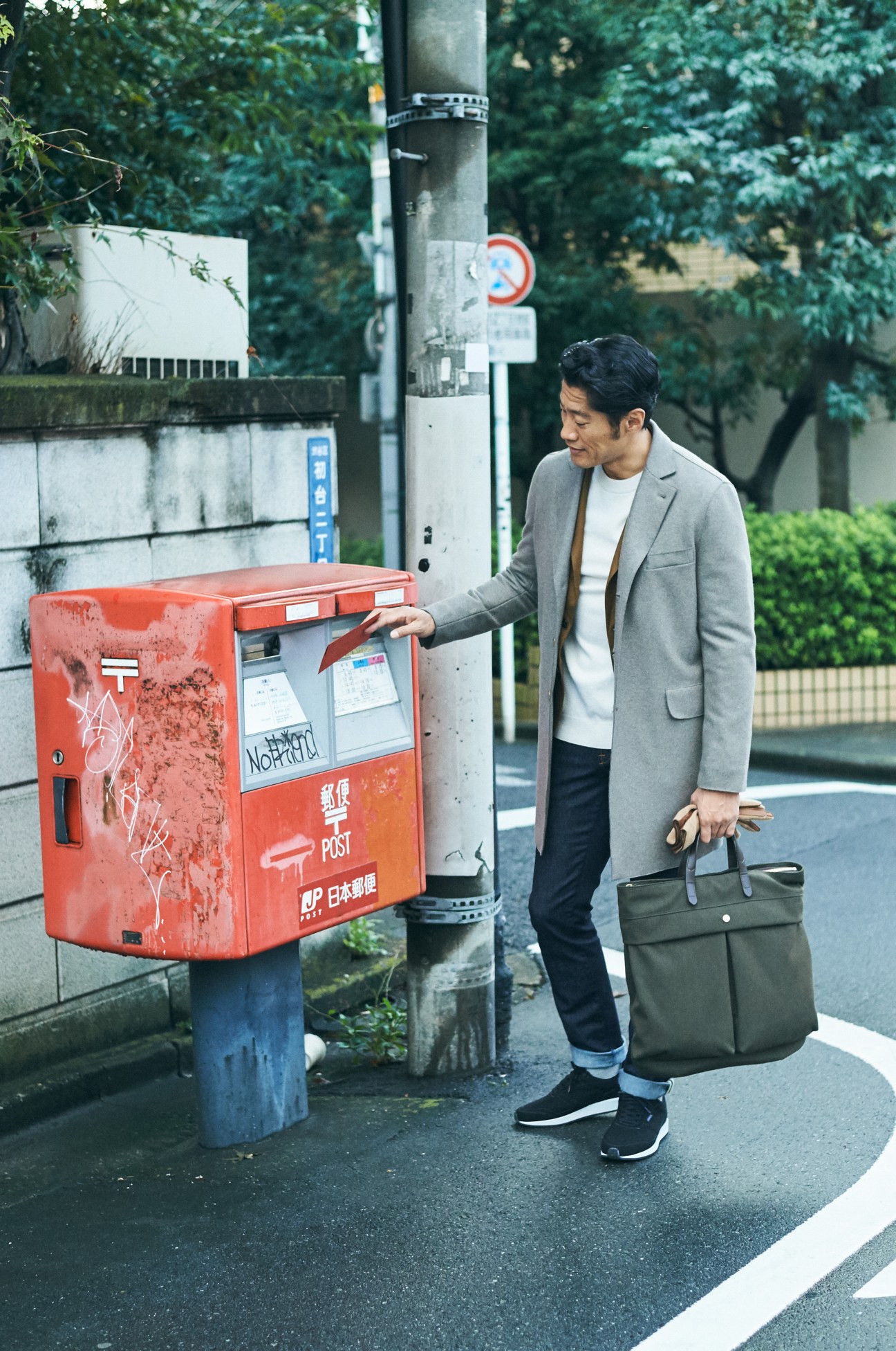 Man in gray coat mailing package at Japanese red postal box on street.