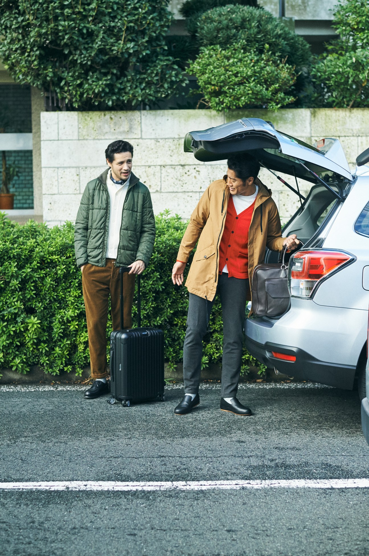 Two men loading luggage into a white SUV with an open tailgate, wearing winter coats and holding a suitcase.