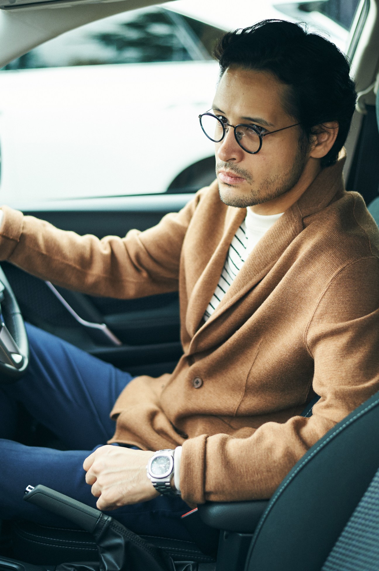 Man wearing camel cardigan and glasses driving a car, watch visible on wrist.