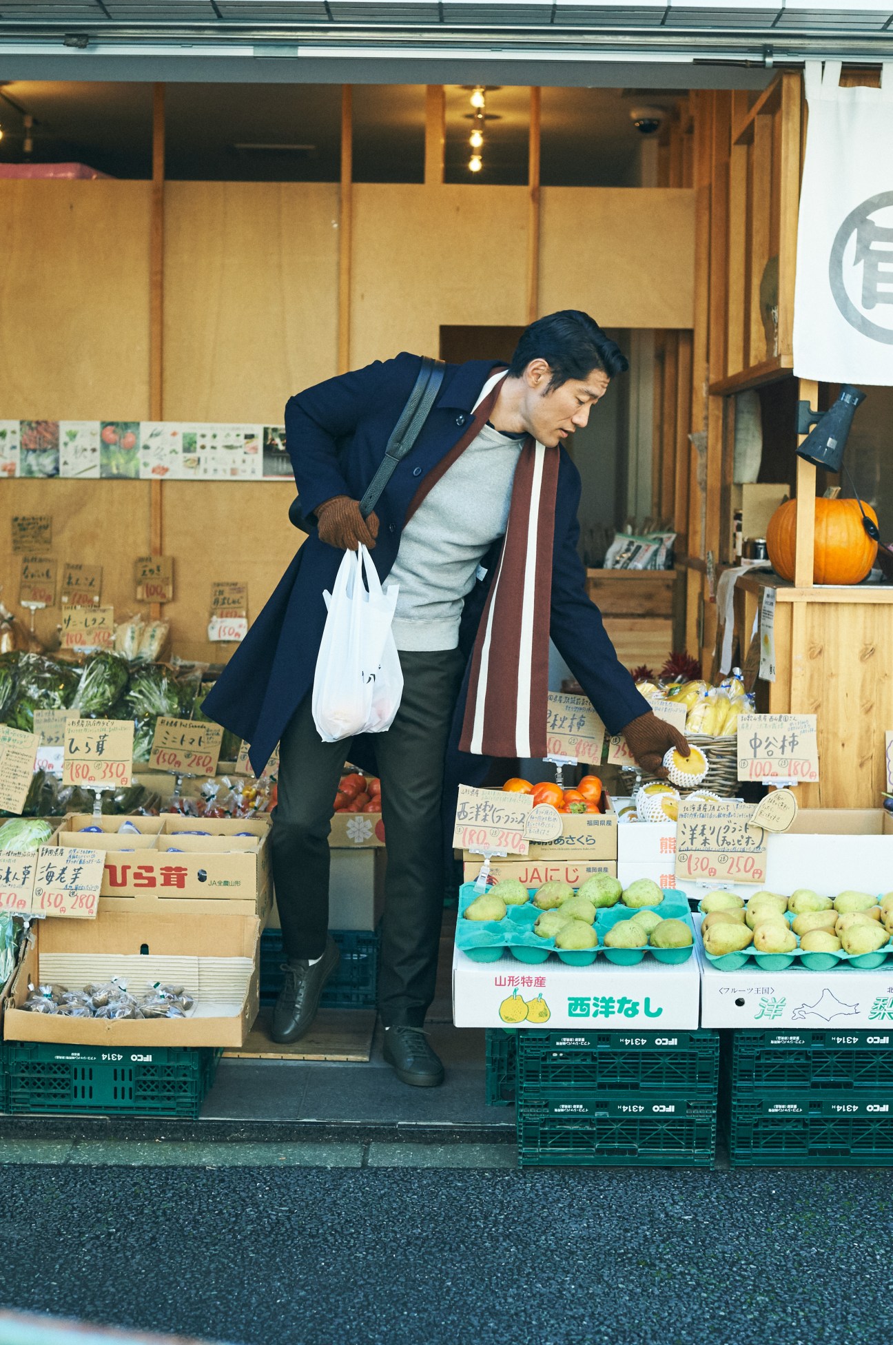 Man in navy coat and burgundy scarf shopping for fruit at a Japanese produce market stall.
